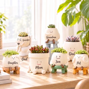 May include: A collection of seven white ceramic planters, each shaped like a small character with dangling legs and a smiling face. The planters are labeled with the words Daughter, Grandpa, Dad, Mom, Grandma, Son, and Pet, and contain various succulents.