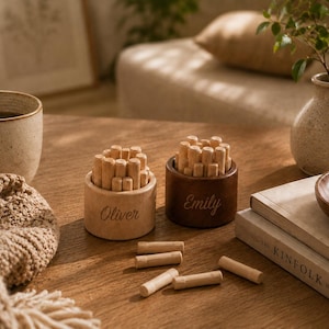 May include: Two cylindrical wooden containers with light and dark brown finishes, each filled with small wooden pegs. The light container is engraved with the name Oliver, and the dark container is engraved with the name Emily. Several loose wooden pegs lie on the surface.