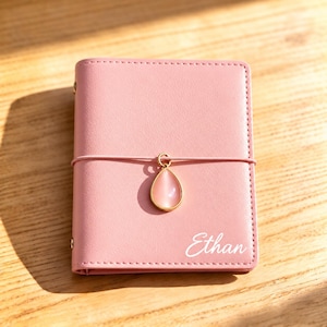 May include: A small, rectangular, light pink notebook with a smooth cover. An elastic band wraps horizontally around the center, holding a gold-rimmed, teardrop-shaped pink stone charm. The name Ethan is printed in white script on the bottom right.