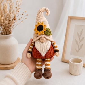 Könnte beinhalten: Eine gehäkelte Zwergenpuppe mit einer beigen, spitzen Mütze, die mit einer gelben Sonnenblume und einem grünen Blatt verziert ist. Der Zwerg hat einen cremefarbenen Bart, einen kürbisförmigen orangefarbenen Körper, gestreifte Arme und Beine in Braun, Weiß und Gelb sowie braune Stiefel.