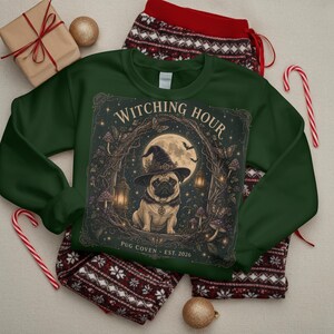 May include: A dark green sweatshirt featuring a graphic of a pug wearing a witch hat, surrounded by a moon, lanterns, and mushrooms. The text reads WITCHING HOUR and PUG COVEN EST. 2026. The sweatshirt is paired with red and white patterned pajama pants.