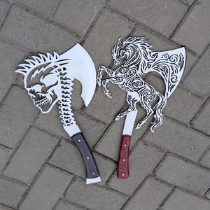 May include: Two metal axes with intricate cutouts. The left axe features a skull and spine design with a dark textured handle. The right axe displays a rearing horse with swirling patterns and a smooth reddish-brown handle.