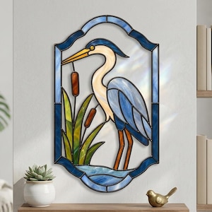 Pode incluir: Um painel de vitral apresentando uma garça azul em pé entre juncos e taboas verdes. A ave tem um pescoço longo, um bico amarelo e plumagem azul e branca. O painel é emoldurado por uma borda azul escura com um formato oval recortado.