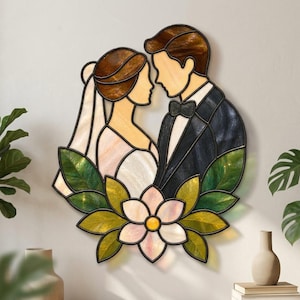 Puede incluir: Un adorno de pared de vidrieras que representa a una novia y un novio frente a frente, enmarcados por hojas verdes y una flor blanca central. La novia lleva un velo y el novio un esmoquin con pajarita, todo realizado en piezas de vidrio.