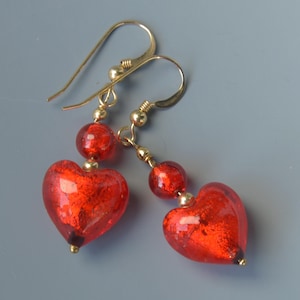May include: A pair of dangling earrings featuring red heart-shaped pendants with a glittery texture. The earrings have gold-colored hooks and small round beads. The heart pendants are a vibrant red color.