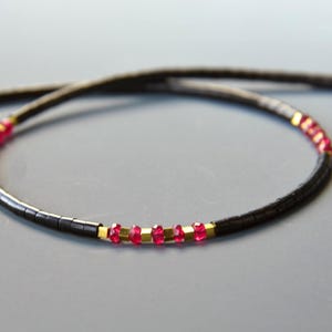 May include: A beaded necklace featuring a long strand of small, cylindrical black beads interspersed with sections of vibrant, translucent red faceted beads and small, square-shaped metallic gold spacers arranged in a repeating pattern.
