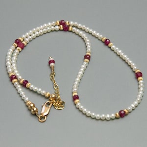 May include: A delicate necklace featuring small, white pearls interspersed with vibrant red gemstones and gold-colored beads. The necklace has a gold-colored clasp and a delicate chain for adjusting the length. The design is elegant and suitable for various occasions.