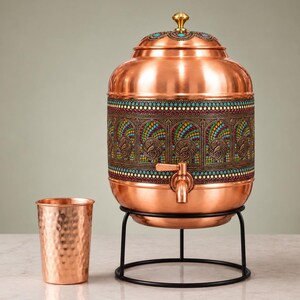May include: A copper water dispenser with a matching hammered copper cup. The dispenser features a central band with intricate peacock motifs and colorful enamel detailing, a brass lid knob, a metal spigot, and a black wire stand for support.
