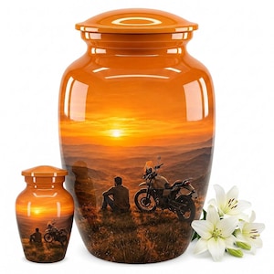 May include: Two orange cremation urns with matching lids, featuring a printed scene of a person sitting on a grassy hill next to a motorcycle at sunset. A cluster of white lily flowers is positioned to the right of the larger urn.