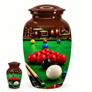 May include: A brown cremation urn featuring a high-resolution wrap-around print of a billiard table with a green felt surface, a rack of red and black balls, a white cue ball, and two wooden pool cues resting on the table surface.