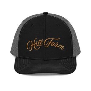 May include: A black trucker hat with a grey mesh back and a solid black front panel. The front features the text Hill Farm embroidered in a gold-colored cursive script. The hat has a curved brim with visible stitching and a black button on top.
