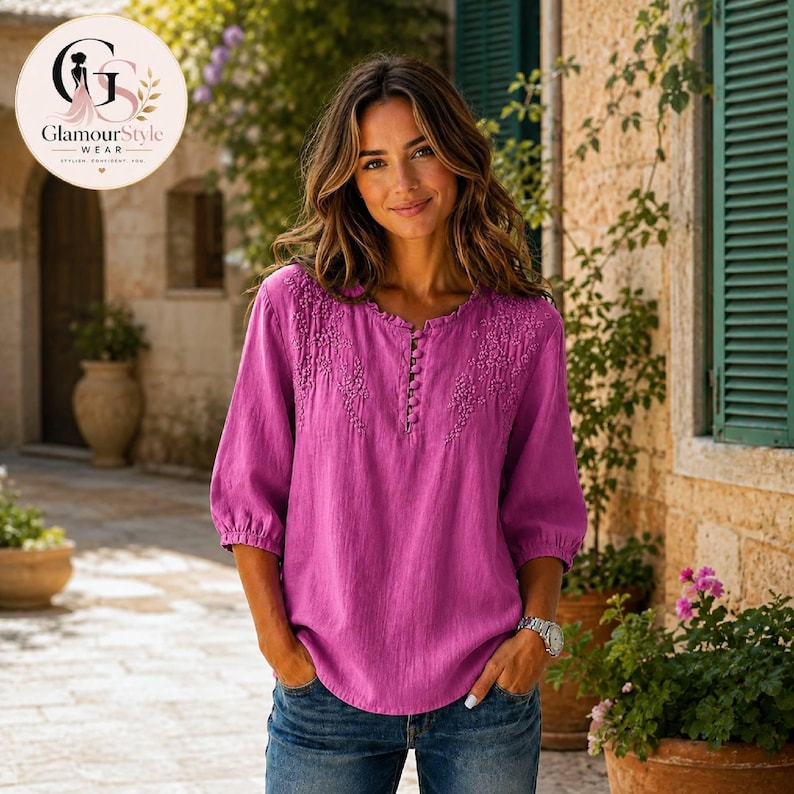 May include: A magenta long-sleeved blouse featuring a round neckline with a vertical row of small buttons and intricate tonal embroidery detailing on the upper chest area. The garment has three-quarter length sleeves with gathered cuffs.