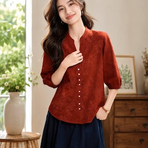 May include: A rust-colored blouse featuring a mandarin collar, three-quarter length sleeves, and a vertical row of small white buttons. The fabric is adorned with an intricate tonal floral embroidery pattern throughout the entire garment.