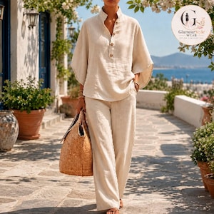 May include: A beige two-piece set consisting of a long-sleeved shirt with a stand collar and four buttons, paired with matching wide-leg trousers. The outfit is made of a textured fabric and is accompanied by a large, woven tan straw tote bag.
