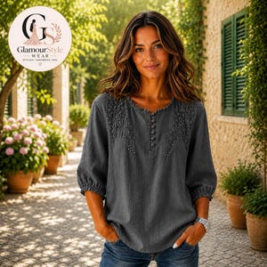 May include: A charcoal gray blouse featuring three-quarter length sleeves with elasticized cuffs, a round neckline, and a vertical row of small buttons. The upper chest area is decorated with tonal floral embroidery, and the fabric has a textured finish.