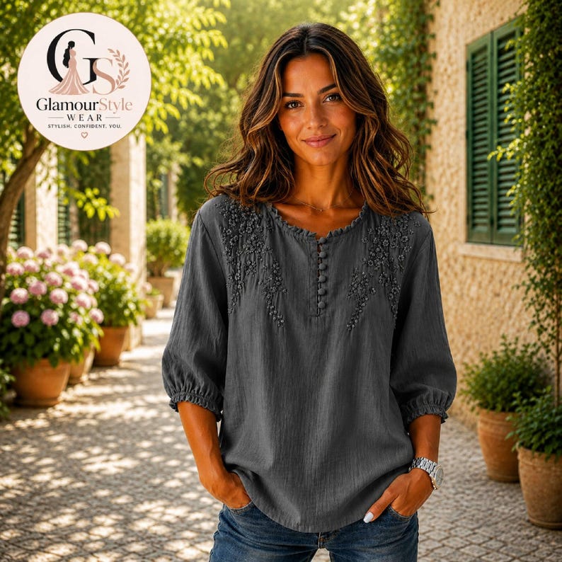 May include: A charcoal gray blouse featuring three-quarter length sleeves with elasticized cuffs, a round neckline, and a vertical row of small buttons. The upper chest area is decorated with tonal floral embroidery, and the fabric has a textured finish.