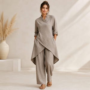 May include: A taupe-colored two-piece set consisting of a long-sleeved tunic with a wrap-style front, a V-neckline, and side pockets, paired with matching wide-leg trousers. The garment features a textured fabric finish and a high-low hemline.