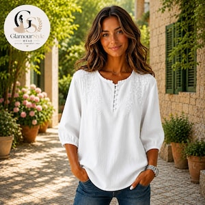 May include: A white long-sleeved blouse featuring a round neckline with a vertical row of small buttons and delicate white floral embroidery on the upper chest area. The garment has a relaxed fit with gathered sleeves and a textured fabric finish.