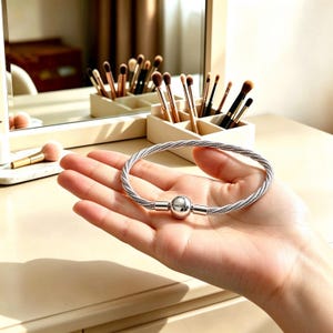 May include: A silver-toned cable bracelet featuring a round, polished clasp engraved with the word PANDORA. The bracelet is constructed from multiple thin, twisted metallic strands that form a flexible, circular band with a secure closure.