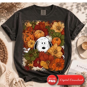 May include: A charcoal gray t-shirt featuring a central graphic of a white dog with black ears surrounded by a vibrant autumn-themed collage of orange pumpkins, red, yellow, and green flowers, and scattered leaves in a rectangular arrangement.