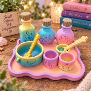 May include: A set of colourful potion-making toys including three bottles with cork stoppers, a mixing bowl with a stirring wand, two small cups, and a spoon on a tray. The items feature pastel gradients and embossed symbols like hearts, moons, and stars.