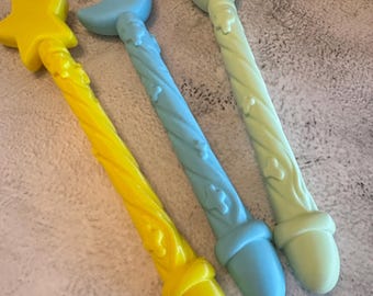 Puede incluir: Tres varitas con mangos trenzados y detalles decorativos. La primera es de color amarillo brillante con una estrella, la segunda es azul claro con una luna creciente y la tercera es verde pálido con una hoja. Cada varita tiene una base redondeada.