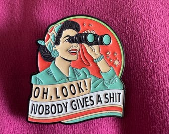 Puede incluir: Un pin de esmalte metálico con una ilustración retro de una persona mirando a través de unos binoculares negros. El fondo es un círculo rojo con pequeñas estrellas blancas. Debajo, una cinta contiene el texto: OH, LOOK! NOBODY GIVES A SHIT.