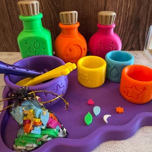 May include: A purple tray holds three colorful potion bottles in green, orange, and pink, three small open cups in yellow, blue, and orange, a purple bowl with a yellow spoon, and a black mesh bag filled with small, colorful plastic shapes.