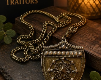 Puede incluir: Un collar con colgante de metal en tono latón que presenta un dije en forma de escudo con un diseño de rosa de los vientos central, adornado con pequeñas perlas blancas en el borde superior y los puntos cardinales, unido a una cadena gruesa.