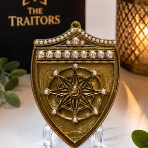 Puede incluir: Un adorno dorado en forma de escudo con un dise&ntilde;o central de estrella de ocho puntas acentuado con peque&ntilde;as perlas blancas. El borde superior est&aacute; decorado con una fila de perlas y la pieza se exhibe sobre un soporte transparente. El texto THE TRAITORS es visible en una caja negra al fondo.