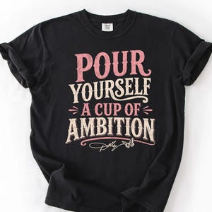 Puede incluir: Una camiseta negra de manga corta con el texto "POUR YOURSELF A CUP OF AMBITION" en una fuente desgastada de color rosa y crema. Debajo del texto hay un peque&ntilde;o gr&aacute;fico de una mariposa y la firma "Dolly" en letra cursiva blanca.