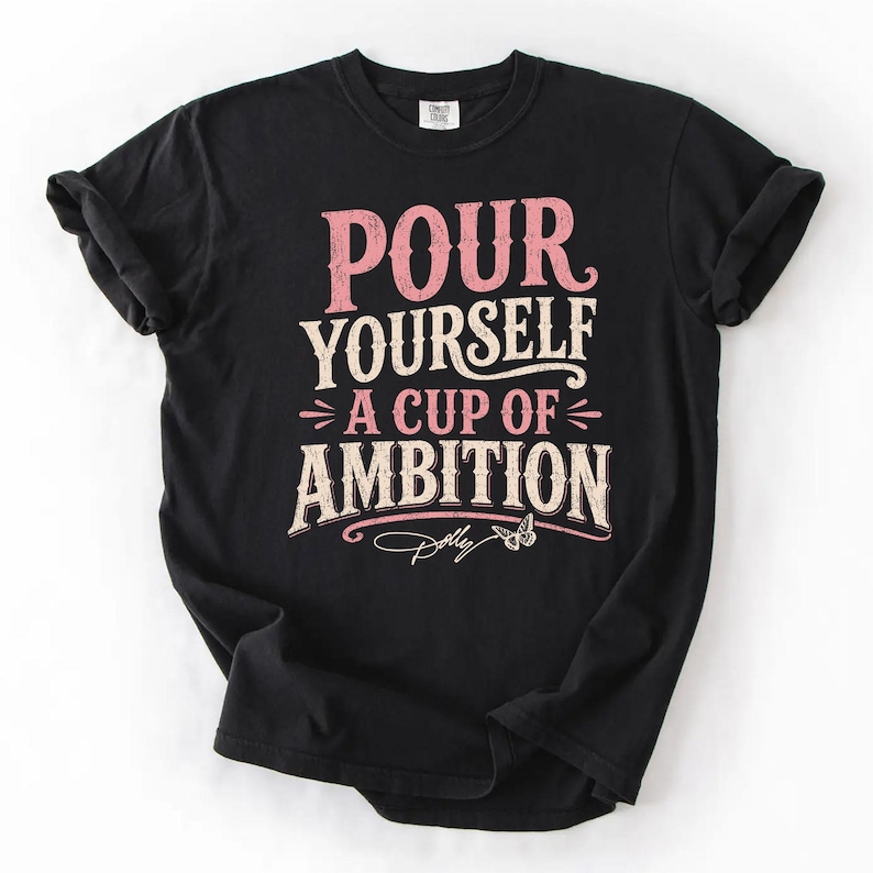Puede incluir: Una camiseta negra de manga corta con el texto "POUR YOURSELF A CUP OF AMBITION" en una fuente desgastada de color rosa y crema. Debajo del texto hay un peque&ntilde;o gr&aacute;fico de una mariposa y la firma "Dolly" en letra cursiva blanca.