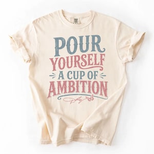 Puede incluir: Una camiseta de manga corta de color crema con el texto "POUR YOURSELF A CUP OF AMBITION" en letras desgastadas de color azul y rosa. Debajo del texto hay un peque&ntilde;o gr&aacute;fico de una mariposa y una firma manuscrita que dice "Dolly".