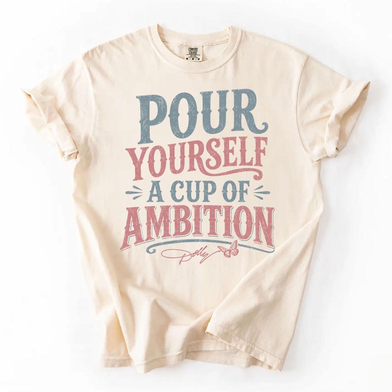 Puede incluir: Una camiseta de manga corta de color crema con el texto "POUR YOURSELF A CUP OF AMBITION" en letras desgastadas de color azul y rosa. Debajo del texto hay un peque&ntilde;o gr&aacute;fico de una mariposa y una firma manuscrita que dice "Dolly".