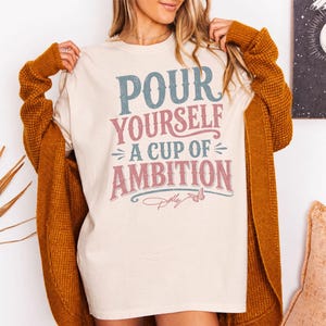 Puede incluir: Una camiseta color crema con el texto "POUR YOURSELF A CUP OF AMBITION" en letras desgastadas azules y rosas, acompa&ntilde;ada de un peque&ntilde;o gr&aacute;fico de mariposa y la firma "Dolly" en letra cursiva rosa, usada bajo un c&aacute;rdigan amarillo mostaza.