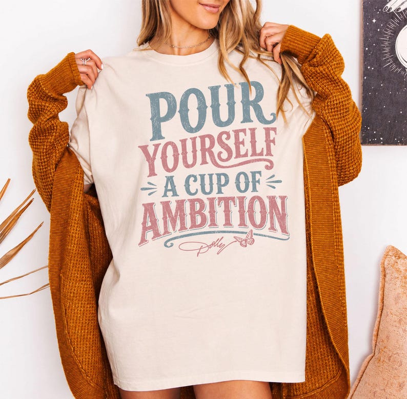 Puede incluir: Una camiseta color crema con el texto "POUR YOURSELF A CUP OF AMBITION" en letras desgastadas azules y rosas, acompa&ntilde;ada de un peque&ntilde;o gr&aacute;fico de mariposa y la firma "Dolly" en letra cursiva rosa, usada bajo un c&aacute;rdigan amarillo mostaza.