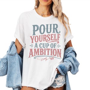 Puede incluir: Una camiseta blanca de manga corta con el texto azul y rosa desgastado "POUR YOURSELF A CUP OF AMBITION", un peque&ntilde;o gr&aacute;fico de mariposa y la firma "Dolly" estampados en el frente, usada con una chaqueta de mezclilla azul claro.