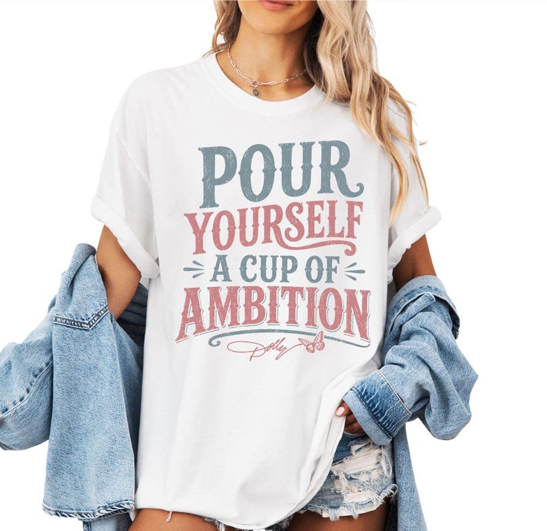 Puede incluir: Una camiseta blanca de manga corta con el texto azul y rosa desgastado "POUR YOURSELF A CUP OF AMBITION", un peque&ntilde;o gr&aacute;fico de mariposa y la firma "Dolly" estampados en el frente, usada con una chaqueta de mezclilla azul claro.