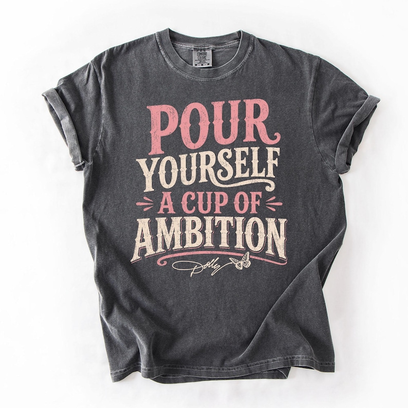 Puede incluir: Una camiseta de manga corta gris carb&oacute;n con el texto en rosa y crema "POUR YOURSELF A CUP OF AMBITION", un peque&ntilde;o gr&aacute;fico de mariposa y la firma "Dolly" estampados en el frente. La camiseta tiene cuello redondo y mangas remangadas.