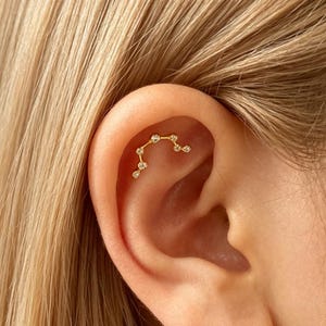 May include: A gold-toned ear cartilage earring featuring a curved, arching design set with six small, clear, round-cut stones. The jewelry is positioned along the upper inner curve of the ear, following the natural contour of the cartilage.