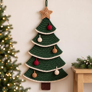 Puede incluir: Un árbol de Navidad de ganchillo para colgar en la pared, de color verde oscuro con una estrella beige en la punta y un tronco marrón. El árbol tiene cinco niveles con bordes festoneados blancos y pequeños adornos de ganchillo en rojo, blanco y beige.