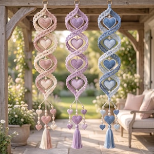 Op de afbeelding: Drie verticale hangende decoraties met gehaakte spiraallinten en hartvormige motieven in roze, paarse en blauwe tinten. Elk stuk bevat kleine witte kralen, extra bungelende hartbedels en een bijpassend garenkwastje.