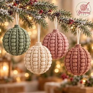 Op de afbeelding: Vier bolvormige gehaakte kerstballen hangend aan een dennentak. Elk ornament heeft een textuurrijk noppenpatroon in saliegroen, crème, oudroze en terracotta, voorzien van een wit koordje om op te hangen en het Yarnora Art logo.