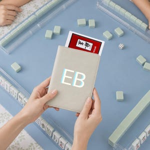 May include: A light gray passport-style holder featuring the letters EB in white and light blue, held over a blue mahjong mat with clear tile racks, scattered light green mahjong tiles, a single die, and a red and white instruction card.
