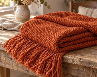 May include: A burnt orange knitted throw blanket featuring a textured stitch pattern and long, thick fringe along the edge. The blanket is folded in layers, showcasing the consistent weave and the deep, warm colour of the yarn throughout the piece.