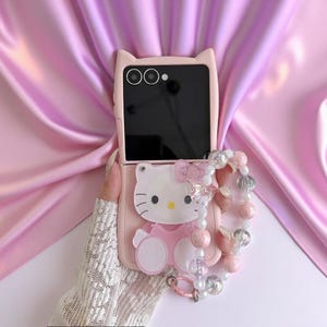 Könnte beinhalten: Eine hellrosa, faltbare Smartphone-Hülle mit Katzenohren-Silhouette am oberen Rand und einer markanten Hello Kitty-Figur auf der Rückseite. Die Hülle enthält ein dekoratives Armband aus verschiedenen rosa, weißen und transparenten Perlen.