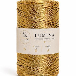 Puede incluir: Un carrete cilíndrico de hilo de algodón metálico dorado con la etiqueta Lumina. La etiqueta especifica un grosor de 1,5 mm, un peso de 200 gramos y una longitud de aproximadamente 285 metros. El hilo tiene una textura brillante y reflectante.