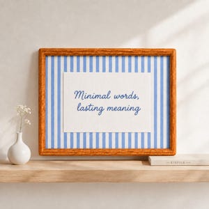 May include: A rectangular wooden frame containing a white print with blue vertical stripes and the blue cursive text Minimal words, lasting meaning. The frame is positioned on a wooden shelf next to a small white vase with a white flower.