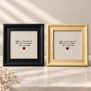 May include: Two square frames, one black and one light wood, each containing a beige fabric insert with black embroidered text that reads: Jeff, we love you to the moon and back, Mom and Dad, accompanied by a small red embroidered heart.