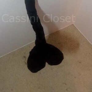 May include: A long, curved, black plush tail, possibly for a costume or cosplay. The tail is thick and fluffy, with a rounded end. The tail is against a white wall and a beige floor.